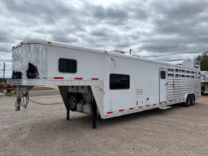 An Exiss Stock Combo livestock trailer with spacious living quarters is parked on gravel under a cloudy sky. The large white trailer features multiple windows, propane tanks, and a sturdy dual-axle design. -Stillwater Trailer Sales