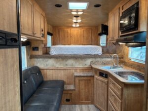 The Exiss Living Quarters Stock Combo RV features wooden cabinetry, a small kitchen with a sink and microwave, a black sofa, and a raised bed in back. Windows on both sides let in natural light, creating a cozy, compact living space. -Stillwater Trailer Sales