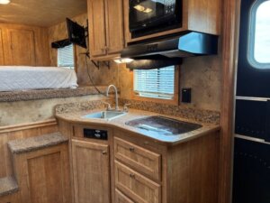 Compact Exiss living quarters RV kitchen with wooden cabinets, sink, stovetop, microwave, and countertop. A small bed with a white quilt is elevated in the background, steps lead up to it. Window blinds and lighting are visible. -Stillwater Trailer Sales