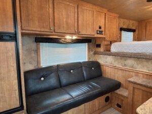 Interior of an Exiss RV with wood cabinets, a black leather couch, a window with blinds, and a raised bed with quilted cover in the living quarters. Steps lead up to the bed and there are built-in countertops for added convenience. -Stillwater Trailer Sales