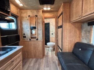 Interior of an Exiss RV with wood cabinetry, a small kitchen area on the left, a black cushioned bench on the right, and a bathroom with a toilet visible through an open door in the back&mdash;perfect living quarters for your next adventure. -Stillwater Trailer Sales