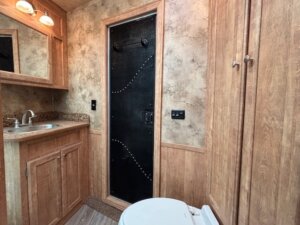 Modern bathroom in the Exiss Living Quarters features wood cabinets, a round sink, mirror with lights above, beige textured walls, a black door with metal accents, and a white toilet in the foreground&mdash;ideal for Stock Combo layouts. -Stillwater Trailer Sales