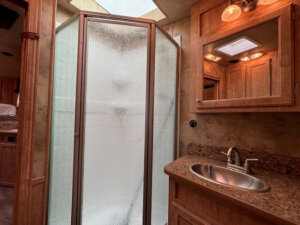 A bathroom in the Exiss Living Quarters features a textured glass shower, stainless steel sink, granite countertop, wooden cabinets, a wall-mounted mirror, and overhead lighting. The space has warm-toned wood finishes and beige walls. Perfect for any Stock Combo setup. -Stillwater Trailer Sales