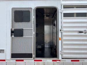 A white Exiss trailer with a screen door and metal door, both open, reveals a dark, empty Stock Combo interior with hooks and equipment inside. Red and white reflective tape lines the lower edge of the trailer. -Stillwater Trailer Sales