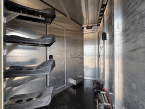The image shows the clean, metallic interior of an Exiss Stock Combo horse trailer, with saddle racks on the left, hooks and storage areas along the walls, and a closed door at the end. Various horse tack and equipment are visible on the floor. -Stillwater Trailer Sales