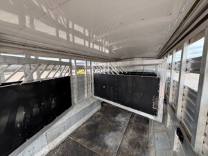 Interior view of an empty Exiss livestock trailer with metal walls, black panels, grated windows, and a dividing gate. Sunlight casts shadows inside the Stock Combo model, while the exterior is visible through the windows. -Stillwater Trailer Sales