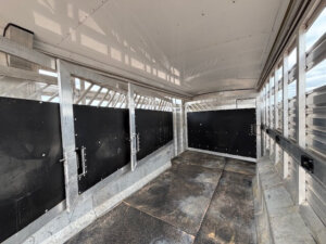 Interior of an empty Exiss Stock Combo trailer with metal walls, black partition gates, and a non-slip floor; sunlight filters through slats on the sides, ready for livestock or Living Quarters conversion. -Stillwater Trailer Sales