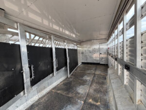 Interior of an empty Exiss Stock Combo trailer with metal walls, open slats for ventilation, black dividers, and a door at the far end. The floor appears worn and well-used. -Stillwater Trailer Sales