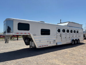 A large, silver Cimarron gooseneck horse trailer with living quarters, five windows, and three axles is parked on gravel under a clear blue sky. -Stillwater Trailer Sales
