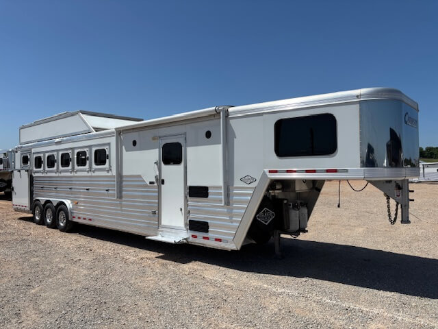 A large silver Cimarron horse trailer with three axles and multiple windows is parked on a gravel lot under a clear blue sky. The 6 horse trailer features a gooseneck hitch, side door, and spacious living quarters. -Stillwater Trailer Sales