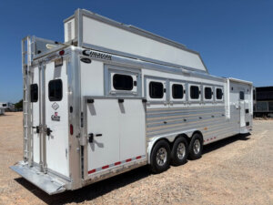 A large, white Cimarron 6 Horse trailer with living quarters, six windows, and three axles is parked on gravel under a clear blue sky. The trailer features a rear door, exterior storage, and prominent Cimarron branding. -Stillwater Trailer Sales
