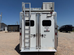 A white Cimarron 6 horse trailer seen from the rear, featuring double doors with windows, a metal ladder on the left, and an air conditioning unit on the roof. The trailer with living quarters is parked on gravel under a clear blue sky. -Stillwater Trailer Sales