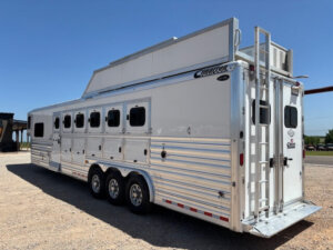 A large silver Cimarron 6 Horse trailer with six windows, triple axles, and a rear ladder is parked on gravel under a clear blue sky. -Stillwater Trailer Sales