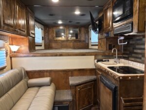 Interior of a Cimarron 6 Horse Living Quarters RV featuring a beige couch, small sink, microwave, TV, wood cabinetry, and a step leading to a raised sleeping area with mirrors and windows with blinds. -Stillwater Trailer Sales