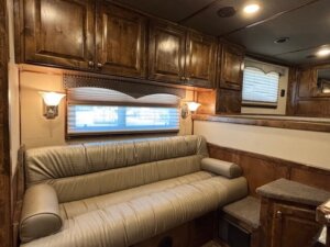 A cozy living quarters horse trailer interior with a beige cushioned sofa, two matching bolster pillows, wooden cabinets above, wall-mounted lights, and a window with blinds and a decorative valance. Light brown paneling and carpeting complete the space. -Stillwater Trailer Sales