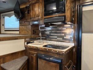 Compact RV kitchen in Cimarron 6 Horse Trailer features wood cabinets, small sink, stovetop, microwave, mini fridge, and modern tile backsplash. TV is mounted upper left; window with blinds sits above a cushioned bench in these cozy living quarters. -Stillwater Trailer Sales