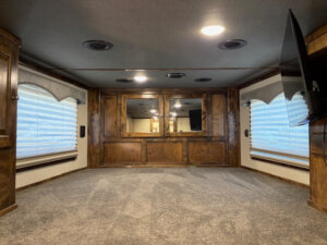 A spacious Cimarron living quarters with gray carpet, wood paneled walls, two large windows with blinds, a wall-mounted TV on the right, and a mirrored section on the far wall under ceiling lights&mdash;perfect for your 6 Horse trailer setup. -Stillwater Trailer Sales