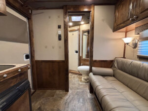 Interior of a Cimarron 6 Horse Trailer featuring 8' Living Quarters with a beige leather sofa, wood cabinets, small kitchen counter, and a bathroom with toilet and mirror. Warm brown and tan tones are highlighted by overhead lighting. -Stillwater Trailer Sales