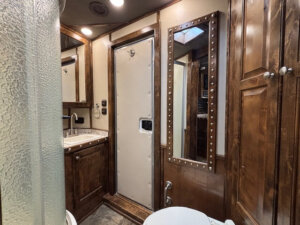 A bathroom in the Cimarron 6 horse trailer&rsquo;s living quarters features wood cabinetry, a sink, wall mirror, tall cabinet, and a door with metal rivets. Warm lighting brightens the space, with part of a glass shower and white toilet visible. -Stillwater Trailer Sales