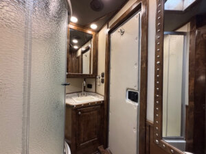Small bathroom in Cimarron 6 Horse Living Quarters, featuring a frosted glass shower on the left, a sink with wooden cabinets and mirror in the center, and a heavy-duty door with metal rivets on the right. Warm lighting brightens the space. -Stillwater Trailer Sales