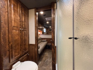 Interior of a Cimarron 6 horse trailer with living quarters: the bathroom in the foreground features a toilet and frosted glass shower, wood-paneled cabinets line the walls, and a cozy seating area is visible in the background. -Stillwater Trailer Sales