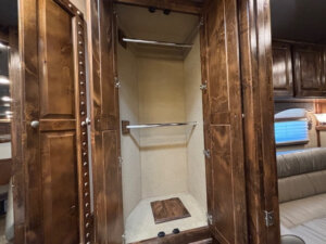 Open wooden wardrobe with two metal hanging rods and an empty beige interior, situated in the 8' living quarters of a Cimarron 6 horse trailer, with brown cabinetry and a beige upholstered sofa visible on the right. -Stillwater Trailer Sales
