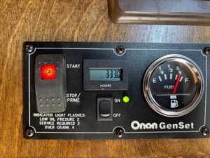 Close-up of a Cimarron Living Quarters Onan GenSet generator control panel showing a red indicator light, digital hour meter at 333.0 hours, fuel gauge, and labeled buttons and switches for start/stop and indicator light codes in a 6 Horse trailer. -Stillwater Trailer Sales