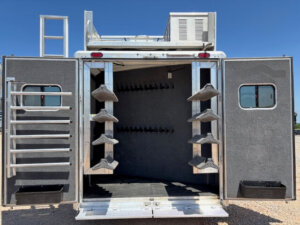 Open rear view of a Cimarron 6 horse trailer with double doors, saddle racks on both sides, bridle hooks inside, a ladder on the left door, rooftop air conditioning unit, and spacious living quarters set against a clear blue sky. -Stillwater Trailer Sales