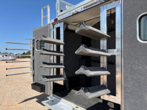 Open Cimarron trailer door revealing a carpeted interior with four angled saddle racks on each side, designed for storing horse saddles; metal bridle hooks are visible in the back. Blue sky and other 6 horse trailers in the background. -Stillwater Trailer Sales
