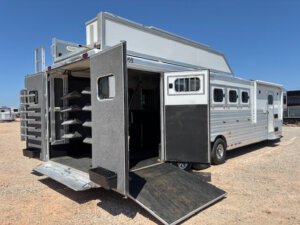 A large Cimarron horse trailer with multiple side windows and open rear doors, revealing a tack room with saddle racks and a ramp extended to the ground, is parked on a gravel lot under a clear blue sky. -Stillwater Trailer Sales