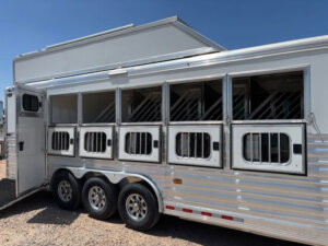 A white Cimarron 6 Horse Trailer with six open windows and metal bars is parked on gravel under a clear blue sky. The trailer features living quarters, three wheels on each side, and an open door at the rear. -Stillwater Trailer Sales