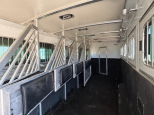 Interior of a Cimarron 6 Horse Trailer with metal dividers creating separate stalls, black and white walls, windows with bars on the right, and a closed door at the far end. -Stillwater Trailer Sales