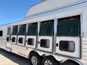 A large silver Cimarron 6 horse trailer with five open windows, each covered by dark mesh screens, is parked outdoors on gravel under a clear blue sky. Designed with living quarters, it offers comfort for both horses and riders. -Stillwater Trailer Sales