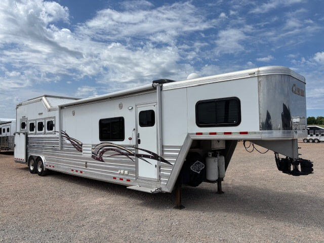 A large Cimarron 4 Horse silver gooseneck trailer with living quarters is parked on gravel under a partly cloudy sky. The trailer features multiple windows, decorative side graphics, and an extended front hitching section. -Stillwater Trailer Sales