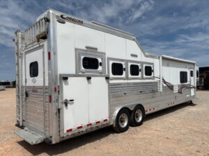 A large white and silver Cimarron 4 horse trailer with living quarters features multiple windows, vents, tandem axles, and graphic stripes along its side. It&rsquo;s parked on gravel under a partly cloudy sky. -Stillwater Trailer Sales