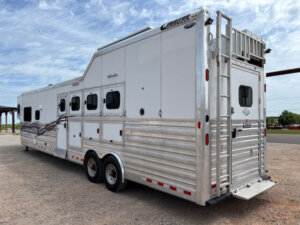 A large white and silver Cimarron 4 horse gooseneck trailer with living quarters, multiple windows, side compartments, a rear ramp, and an exterior ladder is parked on a gravel surface under a partly cloudy sky. -Stillwater Trailer Sales