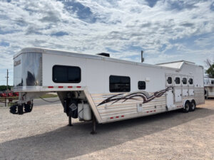 A large white Cimarron 4 Horse Trailer with living quarters, multiple windows, decorative graphics, and three axles is parked on a gravel lot under a partly cloudy sky. -Stillwater Trailer Sales