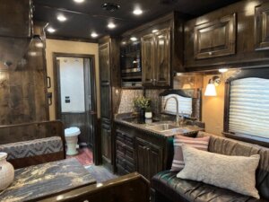 A cozy RV interior inside a Cimarron 4 horse trailer features dark wood cabinets, a small kitchen with sink, stove, and microwave, a cushioned bench with pillows, dining area, and a view of the bathroom in the living quarters. -Stillwater Trailer Sales