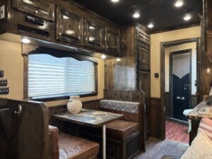 A cozy Cimarron 4 Horse Living Quarters RV interior featuring a dining area with a table and two brown cushioned benches, wood cabinetry, a window with blinds, and a white vase; a bathroom is visible in the background. -Stillwater Trailer Sales