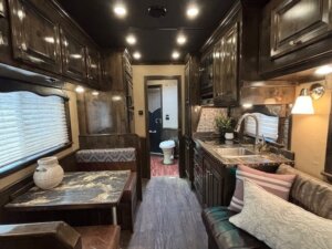Interior of a cozy living quarters horse trailer with a small kitchen, dining area, and seating. Dark wood cabinets, marble-top table with benches, a couch with pillows, and a view of the bathroom in back. Warm lighting throughout. -Stillwater Trailer Sales