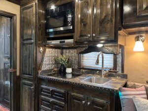 A cozy RV kitchen with dark wood cabinets, a marble countertop, and a stainless steel sink sits within spacious living quarters, featuring a built-in microwave, small potted plant, and cushioned seating area beside a window in this 4 Horse Trailer. -Stillwater Trailer Sales