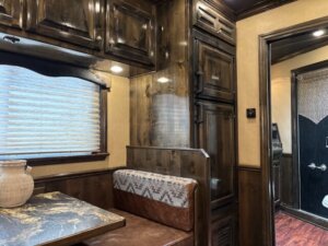 A cozy Cimarron 4 Horse Trailer interior with dark wood cabinetry, a marble-patterned table, brown upholstered bench seating, a decorative vase, and a window with blinds; a door opens to the Living Quarters bathroom in the background. -Stillwater Trailer Sales