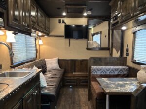 Cozy Living Quarters inside this Cimarron 4 Horse Trailer feature dark wood cabinets, a black sofa, a compact kitchen with a sink, a dining area with patterned bench seating, wall-mounted TV, and windows with blinds&mdash;warm lighting creates an inviting atmosphere. -Stillwater Trailer Sales