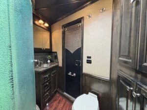 Modern bathroom in Cimarron 4 Horse Trailer living quarters features dark wood cabinets, a white toilet, glass shower door, sink with mirror and countertop, recessed lighting, beige walls with black trim, and wooden flooring. -Stillwater Trailer Sales