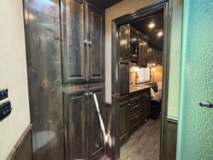 A hallway in a Cimarron 4 Horse Trailer with dark wood cabinets on the left, a frosted glass door on the right, and a view into spacious living quarters and kitchen with matching wood cabinetry ahead. -Stillwater Trailer Sales