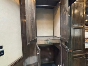 An empty, dark wood cabinet with three open doors reveals shelves and storage space&mdash;perfect for organizing essentials in the living quarters of a Cimarron 4 Horse trailer. The cabinet is built into a wall, next to a section with folded towels. -Stillwater Trailer Sales
