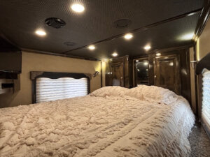 Luxurious living quarters feature a large, textured cream bedspread, wood-paneled cabinets, built-in ceiling lights, and a window with a pleated shade, creating a cozy, elegant atmosphere inside the Cimarron 4 horse side load. -Stillwater Trailer Sales