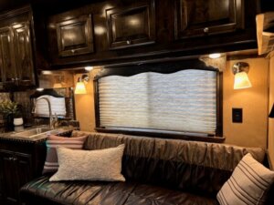 A cozy RV interior inside a Cimarron 4 Horse Trailer features dark wood cabinets, a small sink, and a window with closed blinds. A cushioned sofa with striped and textured pillows sits beneath warm wall lights in the living quarters. -Stillwater Trailer Sales