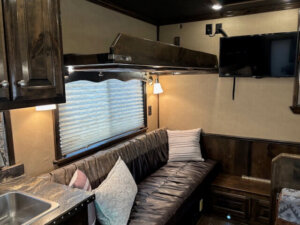 A cozy Cimarron 4 Horse Trailer interior features a brown leather sectional sofa with assorted pillows, a window with closed blinds, a mounted TV, wooden cabinets, and a small sink in the corner. Warm lighting creates a comfortable atmosphere. -Stillwater Trailer Sales
