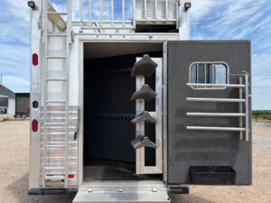 Open tack storage compartment of this Cimarron 4 Horse Trailer with vertical saddle rack, bridle hooks, and open door featuring blanket bars and a small bin. Exterior view shows gravel ground, blue sky, and clouds&mdash;perfect for living quarters setups. -Stillwater Trailer Sales